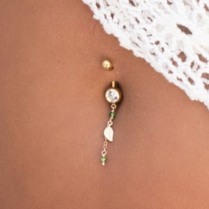 Puede incluir: Un piercing de ombligo dorado con una gema transparente y un pequeño dije de hoja.