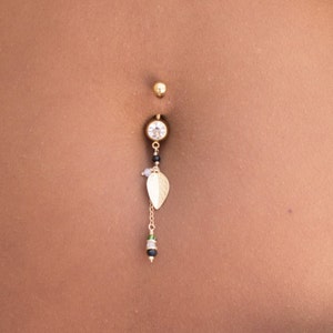 Op de afbeelding: Een close-up van een navelpiercing met een gouden ketting en een klein bladbedeltje. De piercing is op een persoon met een gebruinde huid.