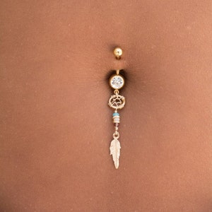 May include: A gold belly button ring with a clear crystal and a dangling feather charm. The charm has a small beaded section with turquoise and brown beads.