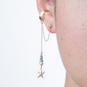 Op de afbeelding: Gouden oormanchet met een hangende ketting en een zeester-bedel. De ketting heeft een parel en turquoise kralen.