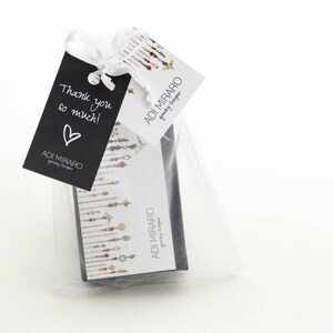May include: A small white gift bag with a black tag that says "Thank you so much!" and a heart. The bag contains a small black box with a colourful design and the text "ADI MIRARO Jewellery Design" on the front.