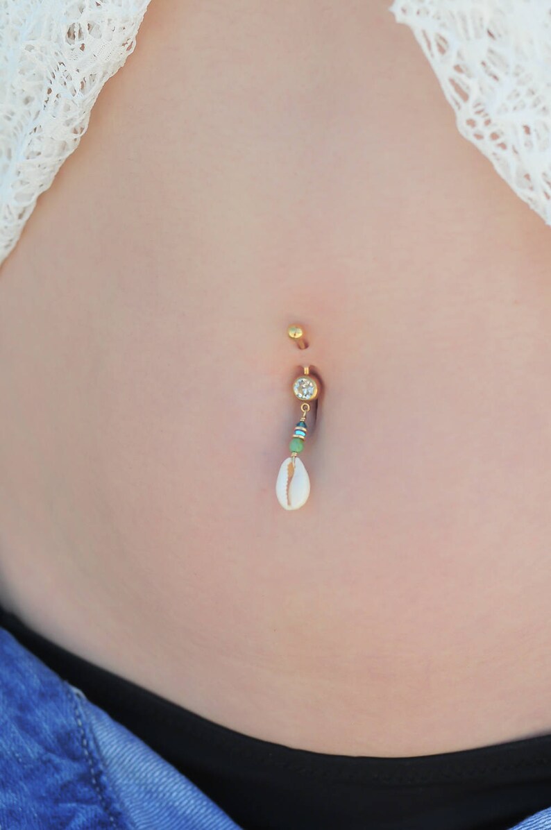 May include: A gold belly button ring with a clear gem and a white seashell charm hanging from it.