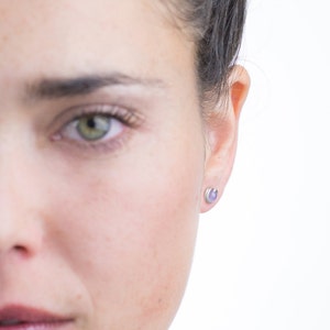 Puede incluir: Primer plano de la cara de una persona, con enfoque en su ojo izquierdo y oreja. Lleva pequeños pendientes redondos de color lavanda.