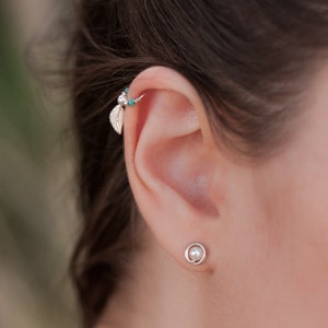 May include: A close-up of a person's ear with two silver earrings. The top earring is a hoop with a small leaf charm and turquoise beads. The bottom earring is a small pearl with a silver ring around it.