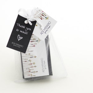 Op de afbeelding: Een kleine witte cadeauzak met een zwart label waarop "Bedankt!" en een wit hart staat. De zak bevat een kleine zwarte doos met een wit label waarop "ADI MIRARO Jewelry Designer" staat.