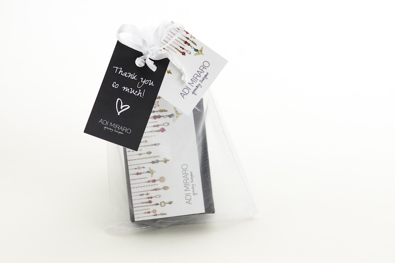 Op de afbeelding: Een kleine witte cadeauzak met een zwart label waarop "Bedankt!" en een wit hart staat. De zak bevat een kleine zwarte doos met een wit label waarop "ADI MIRARO Jewelry Designer" staat.