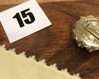 May include: A close-up shot shows a strip of dough with a decorative, scalloped edge, next to a metal pastry wheel. A small white card with the number "15" in black is also visible. The items are on a wooden surface.