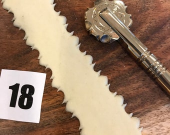 May include: A close-up shot shows a strip of pale dough with a decorative, scalloped edge. A gold-colored, star-shaped pasta cutter rests beside the dough on a wooden surface. A white card with the number 18 is in the lower left corner.