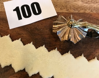 May include: A close-up shot of a wooden cutting board with a decorative pastry cutter and a strip of cut dough. The dough is cut in a zig-zag pattern. A white card with the number "100" in black is also present.
