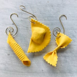 May include: Three yellow pasta-shaped ornaments with gold-colored hooks. The ornaments include a rolled pasta shape, a ravioli shape, and a bow tie pasta shape. The ornaments are displayed on a light gray surface.