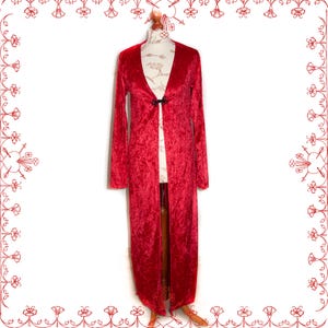 May include: A long, red velvet duster coat with long sleeves and a V-neckline. The coat is fastened with a black clasp at the bust. The garment is displayed on a mannequin.