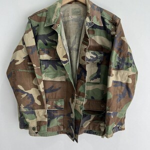 May include: A green, brown and black camouflage jacket with a button closure. The jacket has two large pockets on the front and a smaller pocket on the left sleeve.