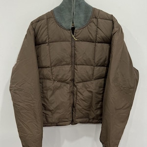 May include: A brown quilted jacket with a gray ribbed collar and cuffs. The jacket has a full zipper closure and a boxy silhouette. The jacket is hanging on a metal hanger against a white background.