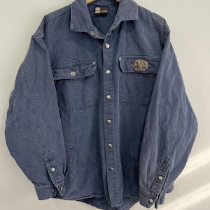 May include: A blue denim jacket with a button-down front, two chest pockets, and two lower pockets. The jacket has silver buttons and a patch on the right chest. The jacket is hanging on a wooden hanger.