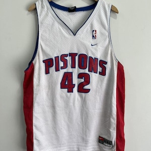 May include: White mesh basketball jersey with the word "PISTONS" in red and blue lettering, and the number "42" in red. The jersey has red side panels, a blue neckline, and a Nike logo.