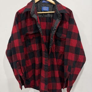 Pode incluir: Uma camisa de manga comprida com botões e um padrão de xadrez vermelho e preto. A camisa tem colarinho, dois bolsos no peito com abas abotoadas e um forro interior preto. Uma etiqueta azul da marca Pendleton é visível no decote interno.