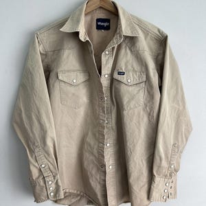 Może przedstawiać: Beżowa koszula Wrangler z długimi rękawami, dwiema kieszeniami na piersi i perłowymi zatrzaskami. Koszula wisi na drewnianym wieszaku na tle białej ściany. Logo Wrangler jest widoczne na kieszeni na piersi i z tyłu kołnierza.