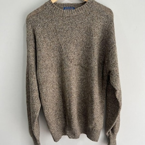 May include: A long-sleeve crew neck sweater in a heathered brown and tan speckled knit pattern. The garment features a ribbed collar, cuffs, and hem, with a blue Pendleton brand label visible at the inner neckline, hanging on a wooden hanger.