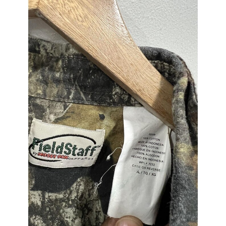 Może przedstawiać: Koszula z guzikami w brązowo-zielono-czarnym wzorze kamuflażu z białą metką, na kt&oacute;rej widnieje napis "FieldStaff by Badger Inc. Apparel" oraz "100% Bawełna Wyprodukowano w Indonezji 100% Bawełna Wyprodukowano w Indonezji 100% Bawełna Wyprodukowano w Indonezji WPL 7232 Instrukcja pielęgnacji na odwrocie AL/TG/XG".