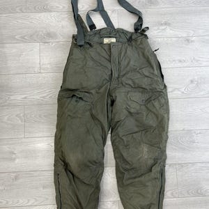 Könnte beinhalten: Olivgrüne, isolierte Latzhose mit verstellbaren grauen Trägern, einem Knopfverschluss vorne, zwei Seitentaschen mit geknöpften Klappen sowie Beinöffnungen mit Reißverschluss und gerippten Bündchen für einen sicheren Sitz bei Outdoor-Aktivitäten.