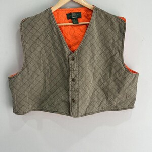 Puede incluir: Un chaleco acolchado con exterior color tostado con patrón de costuras en forma de diamante y forro interior naranja brillante. La prenda tiene cuello en V, cierre frontal con tres botones marrones y una etiqueta de marca verde oscuro.