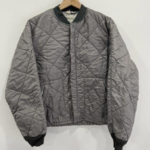May include: A grey quilted bomber jacket with a diamond pattern. The jacket features a dark green ribbed collar and cuffs. It is suspended on a wooden hanger against a white backdrop.
