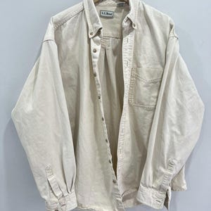 May include: A light beige button-down shirt with long sleeves, a chest pocket, and the brand name "L.L.Bean" on the label. The shirt is hanging on a wooden hanger against a plain background. The shirt has buttoned cuffs and a button-down collar.
