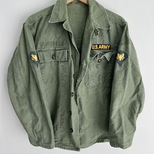 Può includere: Una camicia verde in stile militare con maniche lunghe e chiusura con bottoni. La camicia ha due tasche sul petto e presenta una toppa "U.S. ARMY" sul petto destro e una toppa con un'aquila sulla manica sinistra.