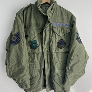 May include: Olive green military-style jacket with multiple pockets and patches. The jacket features the text "US AIR FORCE" on a patch above the chest pocket. Patches with emblems are on the sleeves and chest. The jacket is hanging on a wooden hanger.