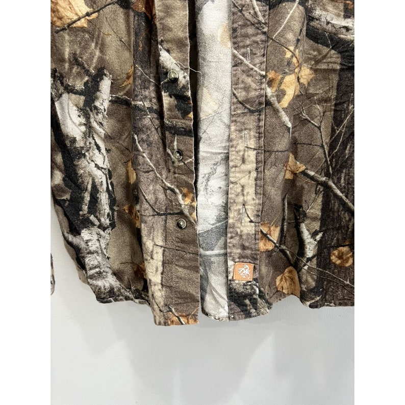 Pu&ograve; includere: Una camicia a maniche lunghe con bottoni di colore mimetico marrone e beige con una piccola toppa marrone in basso a destra.