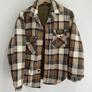 May include: A long-sleeve button-up shirt featuring a brown, cream, and black plaid pattern. The garment includes a pointed collar, two chest pockets with buttoned flaps, and a dark brown fleece lining visible on the interior of the collar.