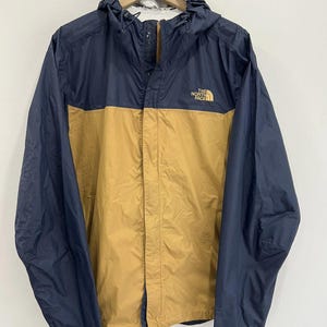 May include: A navy blue and tan hooded jacket with the logo "THE NORTH FACE" on the chest. The jacket has a full zipper closure and a drawstring hood. The sleeves and upper body are navy blue, while the lower body is tan.