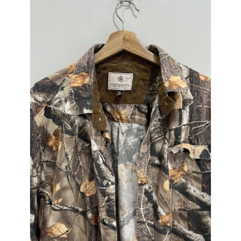 Pu&ograve; includere: Una camicia a maniche lunghe con bottoni in stampa mimetica marrone e beige con colletto in velluto a coste. La camicia ha una toppa sul petto sinistro con il testo "Legendary Whitetails" in bianco.