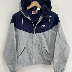 May include: A Nike windbreaker jacket featuring a color-blocked design with a dark navy blue upper section and a light gray lower section. The jacket includes a hood with white drawstrings, a full-length front zipper, and two zippered side pockets.
