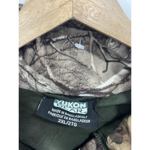 May include: A green and brown camouflage fleece jacket with a zipper closure. The jacket has a label that reads "Yukon Gear" and "Made in Bangladesh/Fabrique' en Bangladesh" and "2XL/2TG".