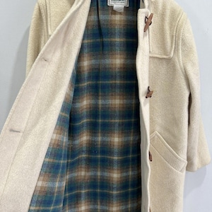 May include: Cream-colored hooded duffle coat with wooden toggles and a plaid lining. The coat features a classic design with a hood, front pockets, and a button closure. The interior lining is a blue, green, and brown plaid pattern. The L.L.Bean label is visible.