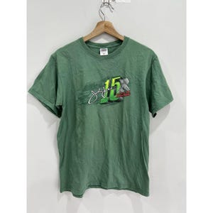 May include: Green short-sleeved t-shirt with a graphic of John Force's signature and the number 15. The graphic also includes the text "15x NASCAR Champion".