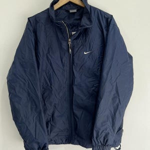 May include: A navy blue windbreaker jacket featuring a full-length front zipper, a high collar, and long sleeves with elastic cuffs. A small white Nike swoosh logo is embroidered on the upper left chest. The jacket has a lightweight, crinkled texture.
