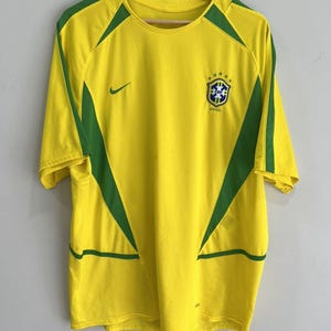 Puede incluir: Camiseta de fútbol amarilla y verde de Brasil con el logo de Nike y el escudo de la selección brasileña. La camiseta tiene detalles verdes en los hombros y los laterales. La camiseta está colgada en una percha de madera.