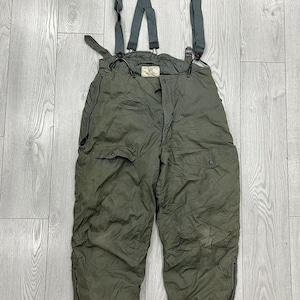 Könnte beinhalten: Olivgrüne, isolierte Schneehose mit angebrachten Hosenträgern. Die Hose hat zwei Vordertaschen, Reißverschlüsse an den Knöcheln und ein Etikett an der Taille. Die Hosenträger sind verstellbar und werden mit Clips befestigt. Aus wasserabweisendem Material.