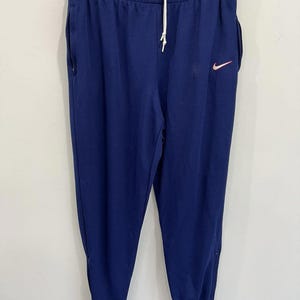 May include: Navy blue sweatpants with a white drawstring and a small red Nike logo. The pants have side pockets and white stripes at the ankles. The pants are hanging on a black hanger.