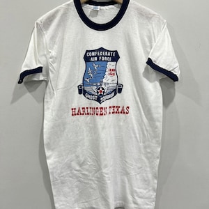 May include: White t-shirt with navy blue trim. The front displays a graphic of the "Confederate Air Force Ghost Squadron Harlingen Texas." The graphic includes a shield with airplanes and the years 1939-1945.