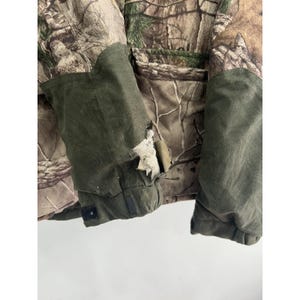 May include: A green and brown camouflage jacket with a torn area on the left sleeve. The jacket has a zipper closure and a hood.