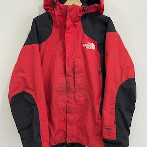Puede incluir: Chaqueta con capucha roja y negra con el texto "THE NORTH FACE" en el pecho. La chaqueta tiene paneles negros en los hombros y las mangas, y una etiqueta "GORE-TEX" en la manga. La chaqueta está colgada en una percha.