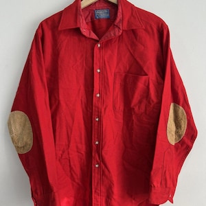 Pode incluir: Uma camisa vermelha brilhante de manga comprida com gola e fecho de botões. A camisa apresenta cotoveleiras bege e um bolso no peito. A camisa está pendurada em um cabide de madeira contra uma parede branca.