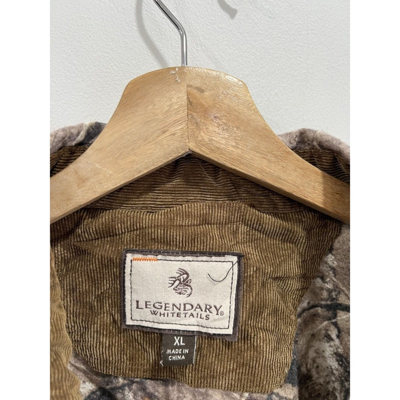 Pu&ograve; includere: Una giacca di velluto a coste marrone con motivo mimetico. La giacca ha un'etichetta che dice "Legendary Whitetails" e "XL Made in China".