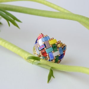 May include: A multi-coloured ring with a gold band, featuring a woven design. The ring is made up of small, rectangular beads in shades of red, blue, yellow, and green. It rests on green stems against a white background.