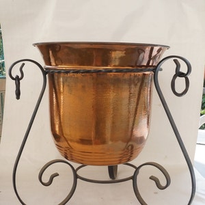 May include: A hammered copper planter with a flared rim, supported by a wrought iron stand. The stand features decorative scrollwork and a twisted metal band around the planter's midsection. The copper has a warm, reflective finish.
