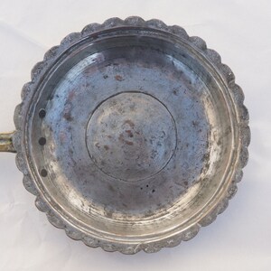 May include: A vintage copper pan with a brass handle and a scalloped edge. The pan has a circular design with a small hole in the center.