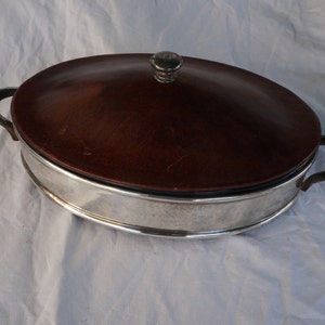 Peut inclure: Un plat de service ovale en argent avec un couvercle en bois brun et deux poignées. Le couvercle a un petit bouton sur le dessus.